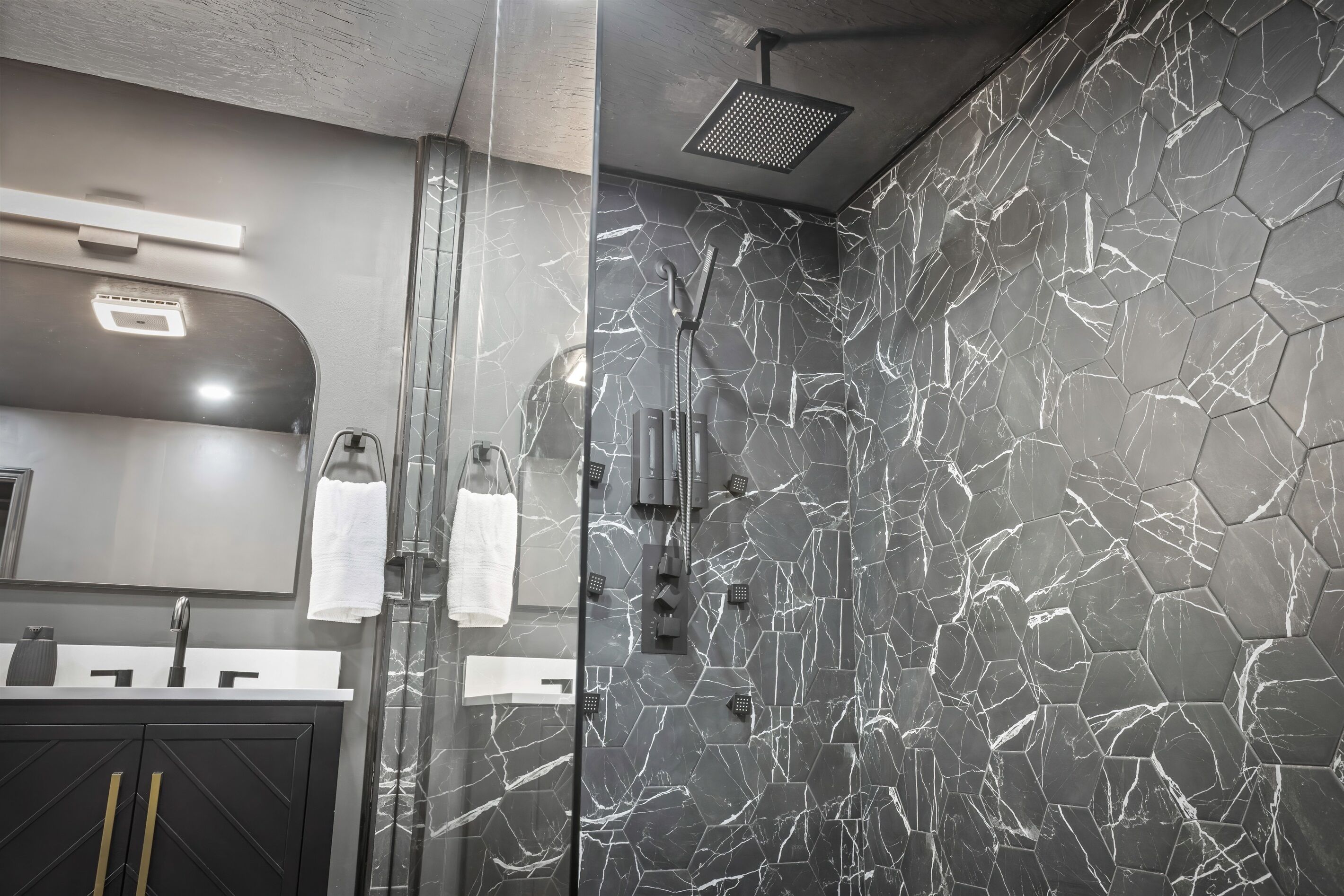 A modern bathroom with a glass shower enclosure, a black marble wall with white veins, a black cabinet with gold handles, and a white towel hanging on the wall.