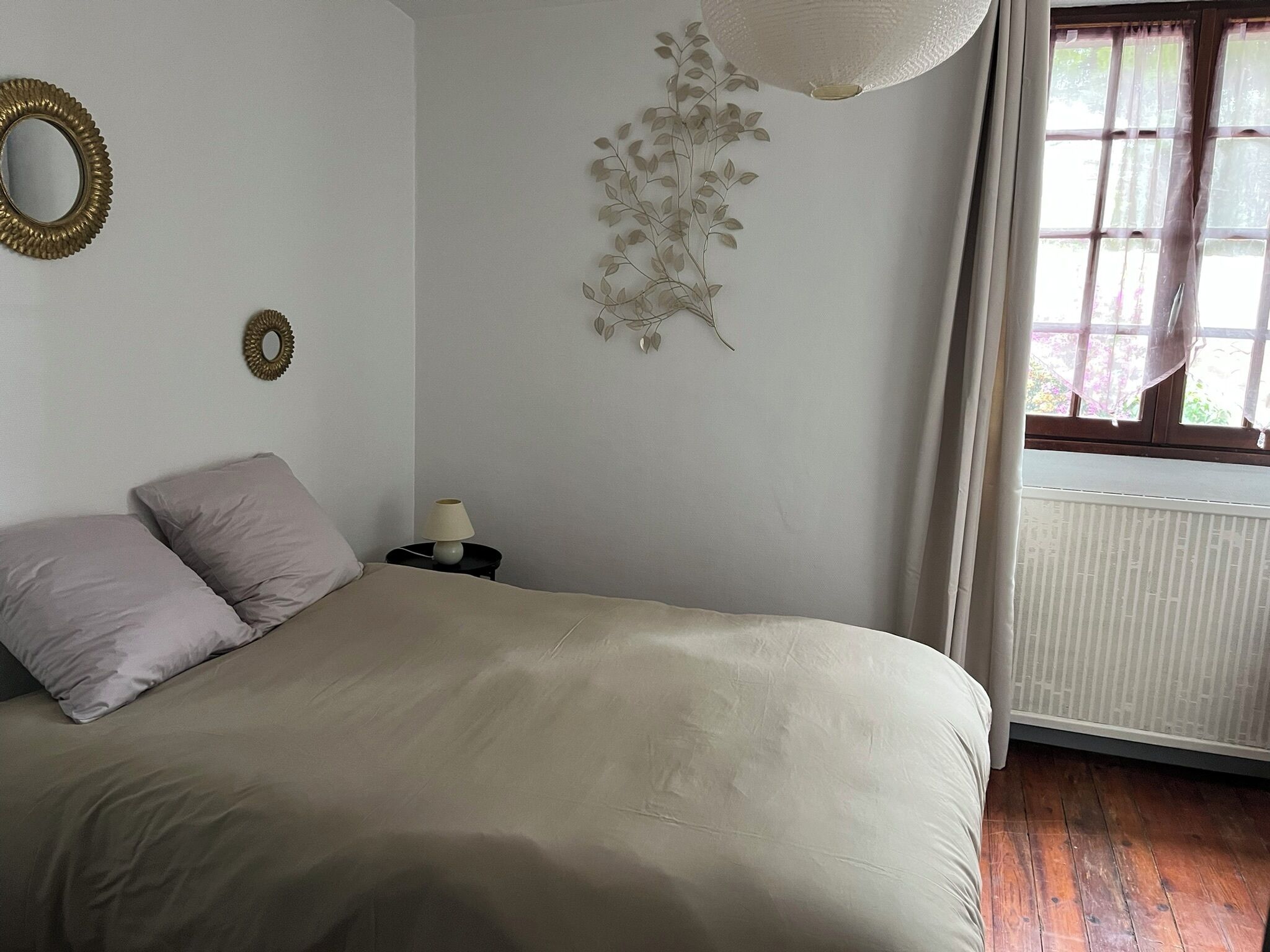 Ein Schlafzimmer mit einem Bett, Kissen, einem Nachttisch mit Lampe, einem Wandspiegel und einem Fenster mit Vorhängen.