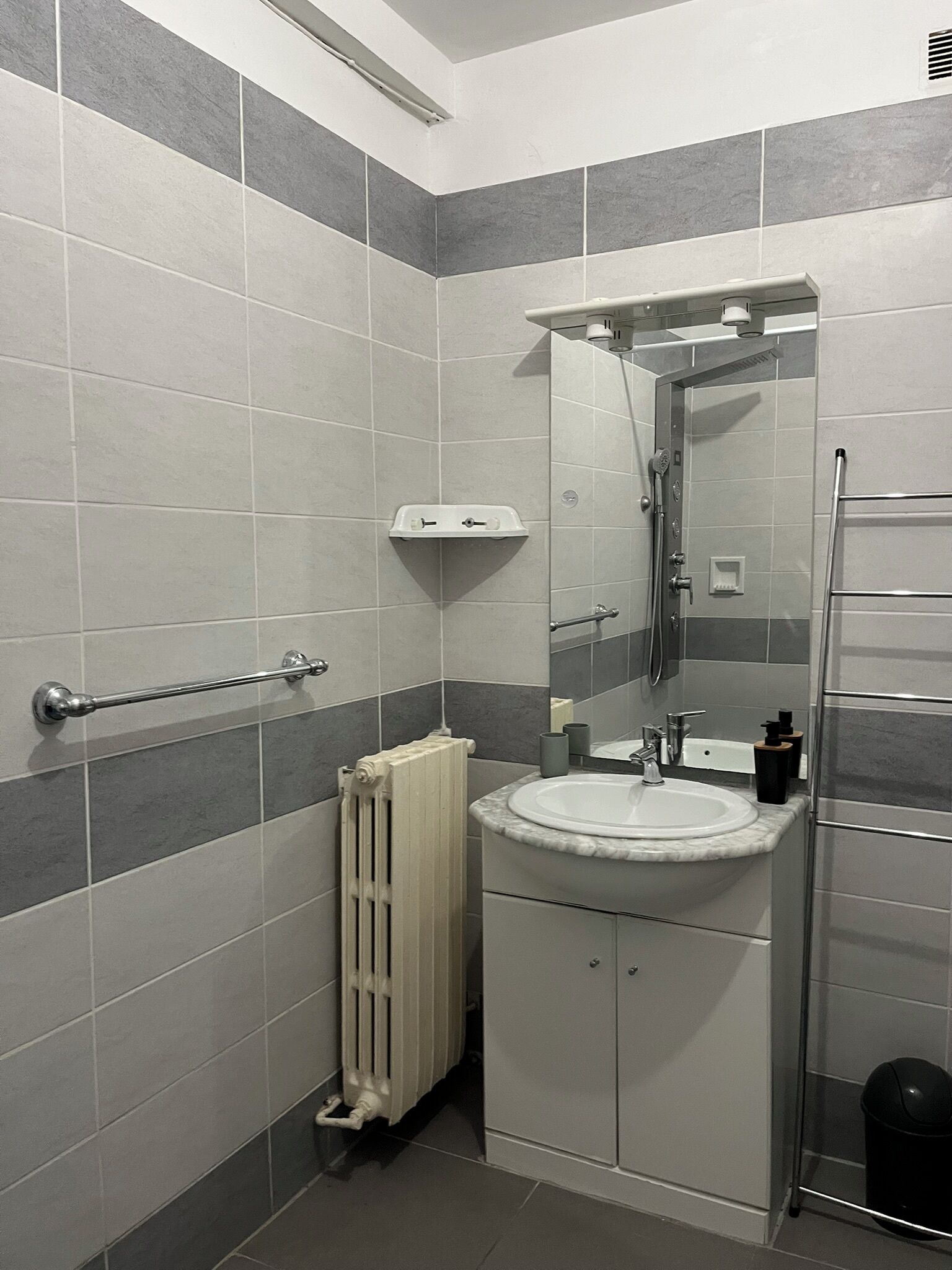 Une salle de bain moderne avec des carreaux gris et blancs, un lavabo blanc, un miroir, un porte-serviettes et un radiateur.