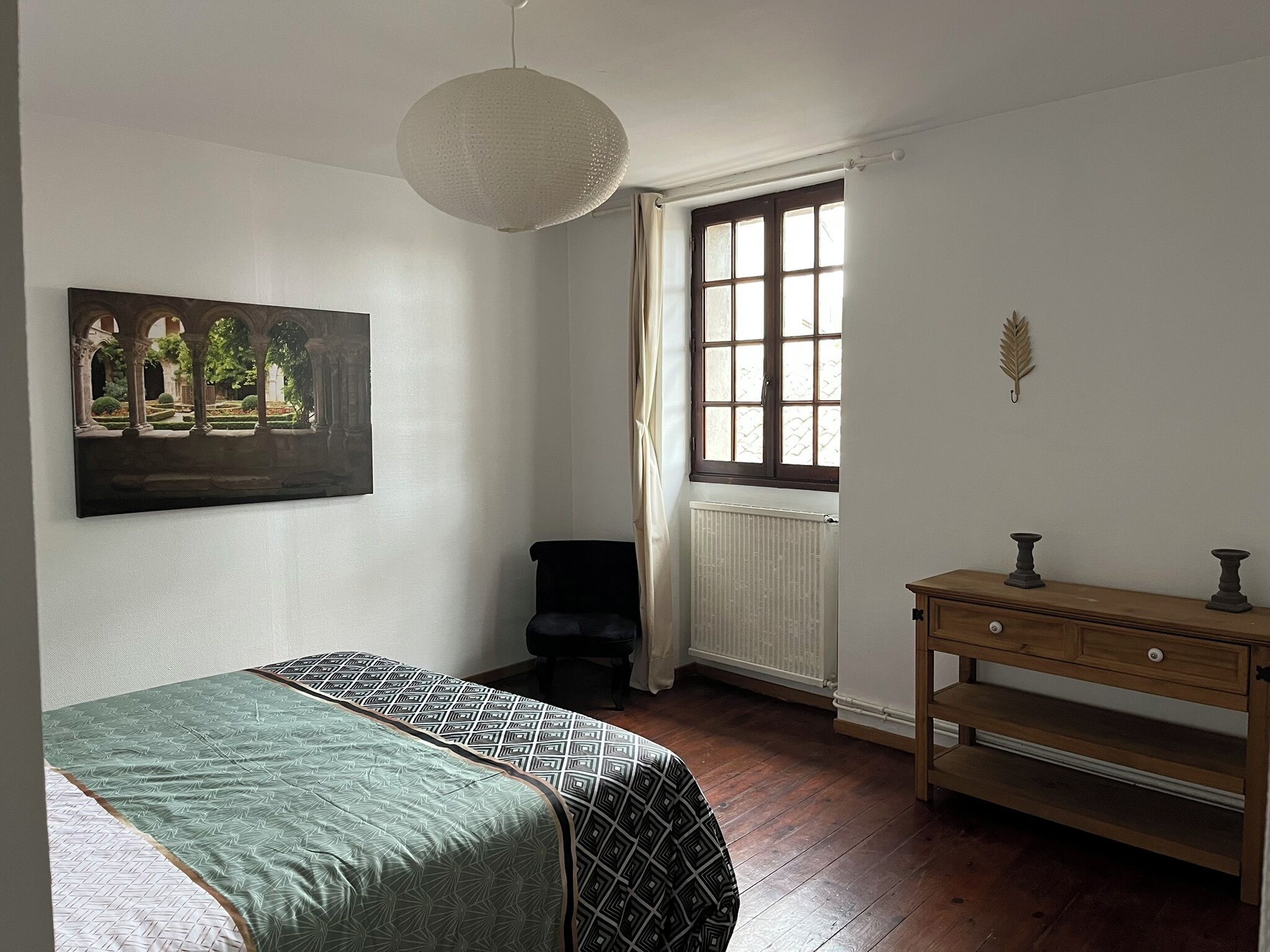 A bedroom with a bed, a window with curtains, a radiator, a wooden dresser, and a painting on the wall.