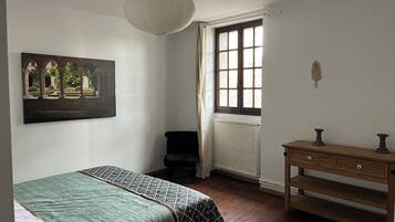 A bedroom with a bed, a window with curtains, a radiator, a wooden dresser, and a painting on the wall.