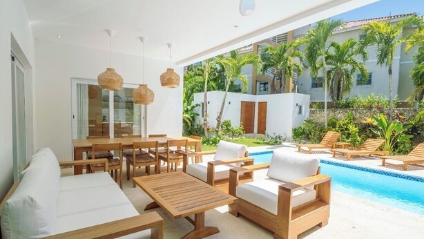 Uma área externa moderna com piscina, móveis de madeira e vista para um prédio branco e palmeiras.