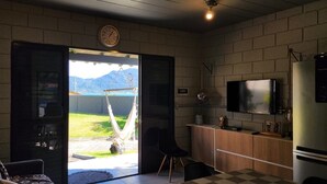 Una habitación con un gran ventanal que da a un balcón, un televisor en la pared, un refrigerador y un pequeño armario de madera.