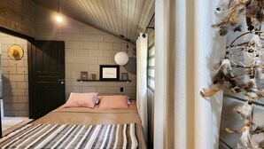 Un dormitorio con cama, almohadas, un estante en la pared con cuadros y una lámpara colgante, además de un atrapasueños.