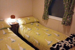 A small room with two single beds, a nightstand with a lamp, and a window with floral curtains. - The Static Cowavan Towyn (Abergele)
