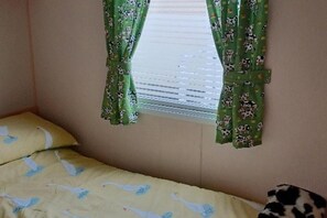 A bedroom with a bed, a window with green curtains, and a pillow with a pattern. - The Static Cowavan Towyn (Abergele)