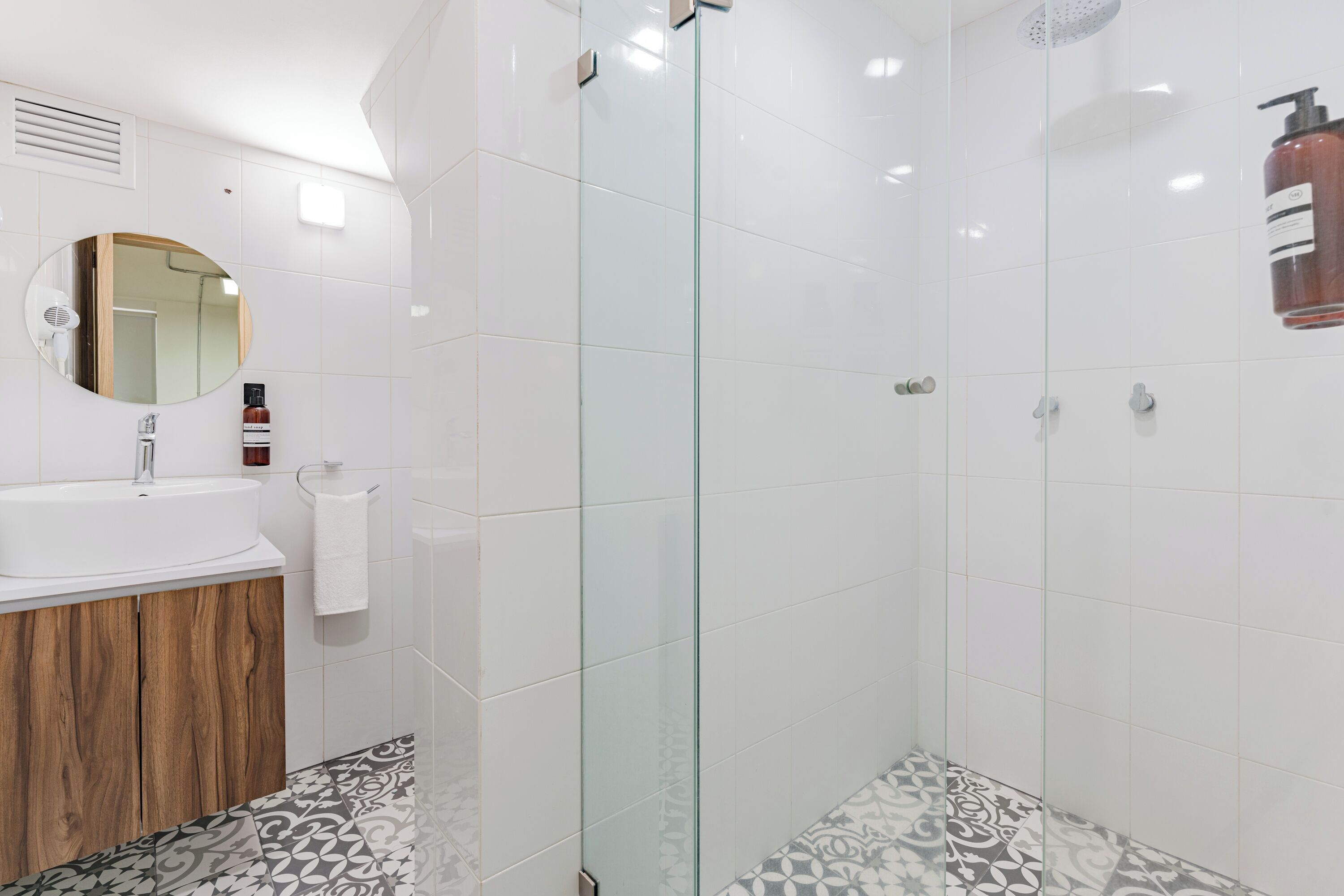 A modern bathroom with a glass shower enclosure, a wooden vanity, and patterned floor tiles.