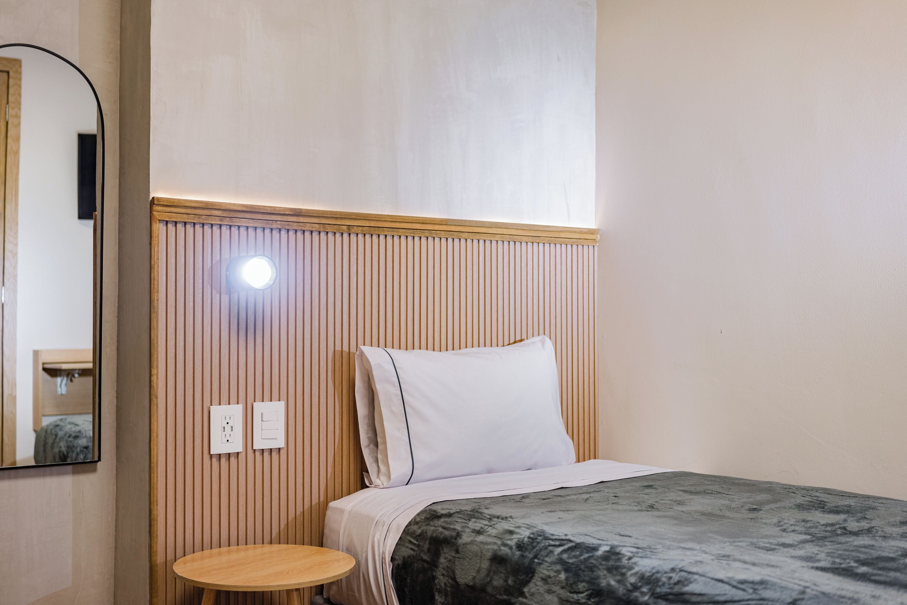 A hotel room with a bed, a round wooden table, a mirror, and a wall-mounted light.