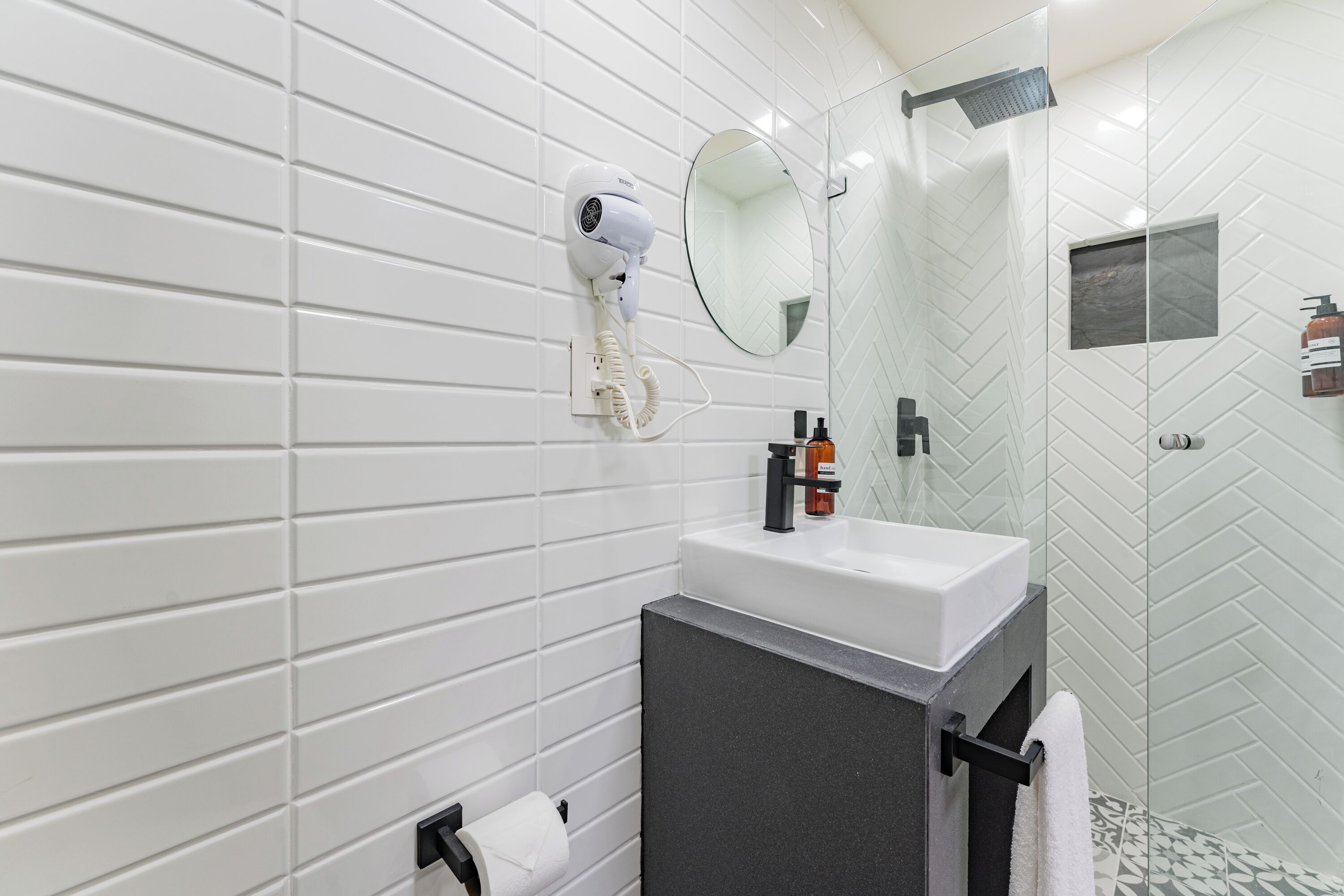 A modern bathroom with white tiled walls, a round mirror, a white sink, and a glass shower enclosure.