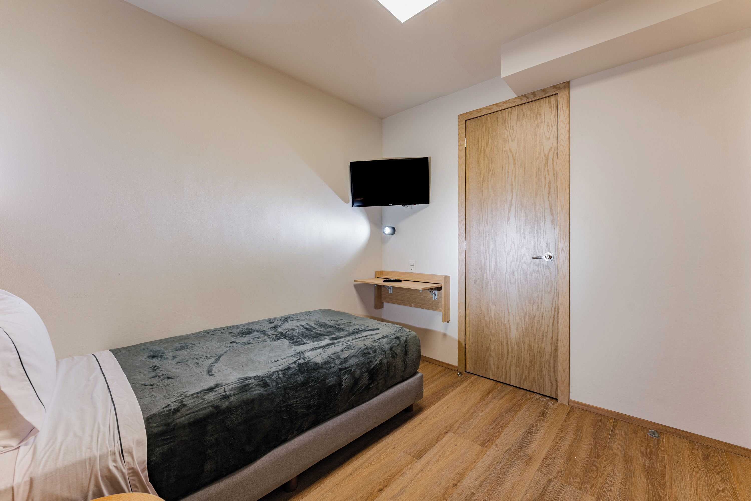 A small bedroom with a bed, a TV mounted on the wall, a wooden door, and a small shelf.