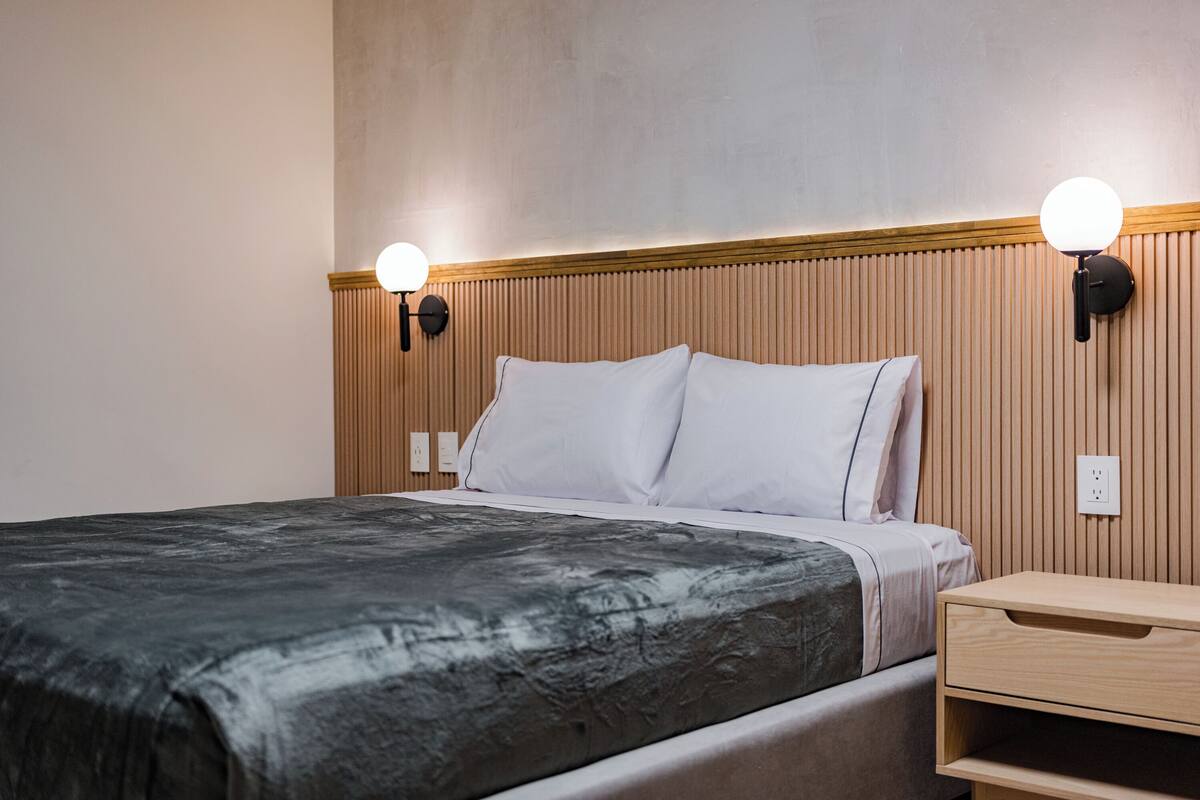 Ein ordentlich bezogenes Bett mit dunkler Decke, ein Nachttisch mit Schublade und zwei Wandleuchten.