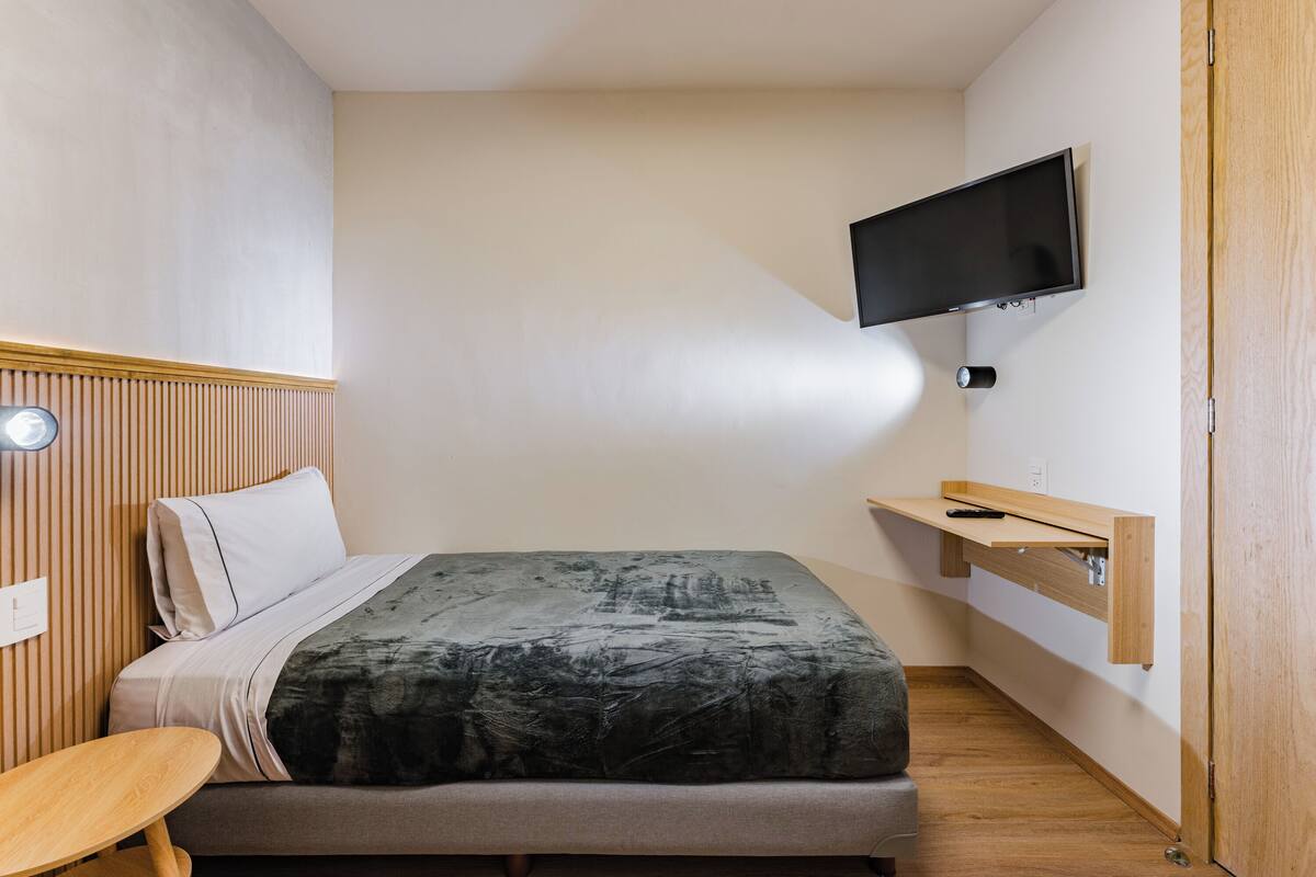 Een kleine slaapkamer met een bed, een tafeltje, een wandtelevisie en een houten deur.