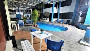 An indoor swimming pool with blue chairs and a table, surrounded by stone tiles and potted plants.