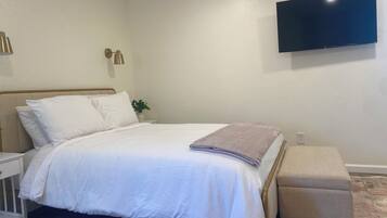 A neatly made bed with white linens, a bedside table with a lamp, and a flat-screen TV mounted on the wall.