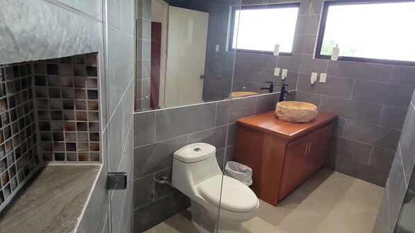 A bathroom with a toilet, a sink with a stone basin, a mirror, and a tiled wall with a window.