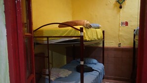 Habitación tipo dormitorio con dos literas, una con cubrecama amarillo y la otra con cubrecama azul. Se observa un estante con ropa doblada y una bolsa negra en la litera inferior.