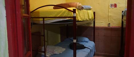 A dormitory room with two bunk beds, one with a yellow bedspread and the other with a blue bedspread. There is a small shelf with folded clothes and a black bag on the lower bunk.
