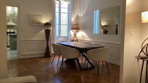 Ein Essbereich mit Holzdielenparkett, einem rechteckigen Tisch und vier weißen Stühlen. Eine hohe Stehlampe und ein wandmontierter Spiegel sind vorhanden. Durch ein mit Vorhängen drapiertes Fenster fällt natürliches Licht herein.