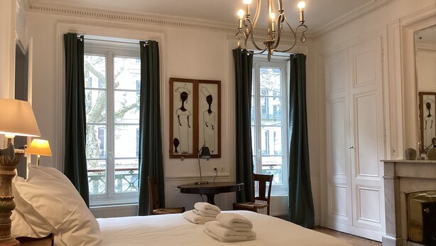 Ein Hotelzimmer mit einem großen Bett, einem Schreibtisch mit Lampe, einem Sessel, einem Kamin und zwei Fenstern mit Vorhängen.