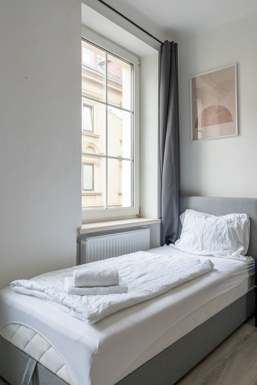 Una cama individual con cabecera gris, ropa de cama blanca y ventana con vistas a edificios.