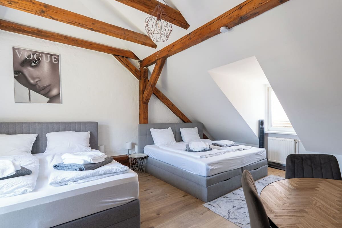 A modern loft bedroom with exposed wooden beams, two beds, a dining table, and a large window providing natural light.