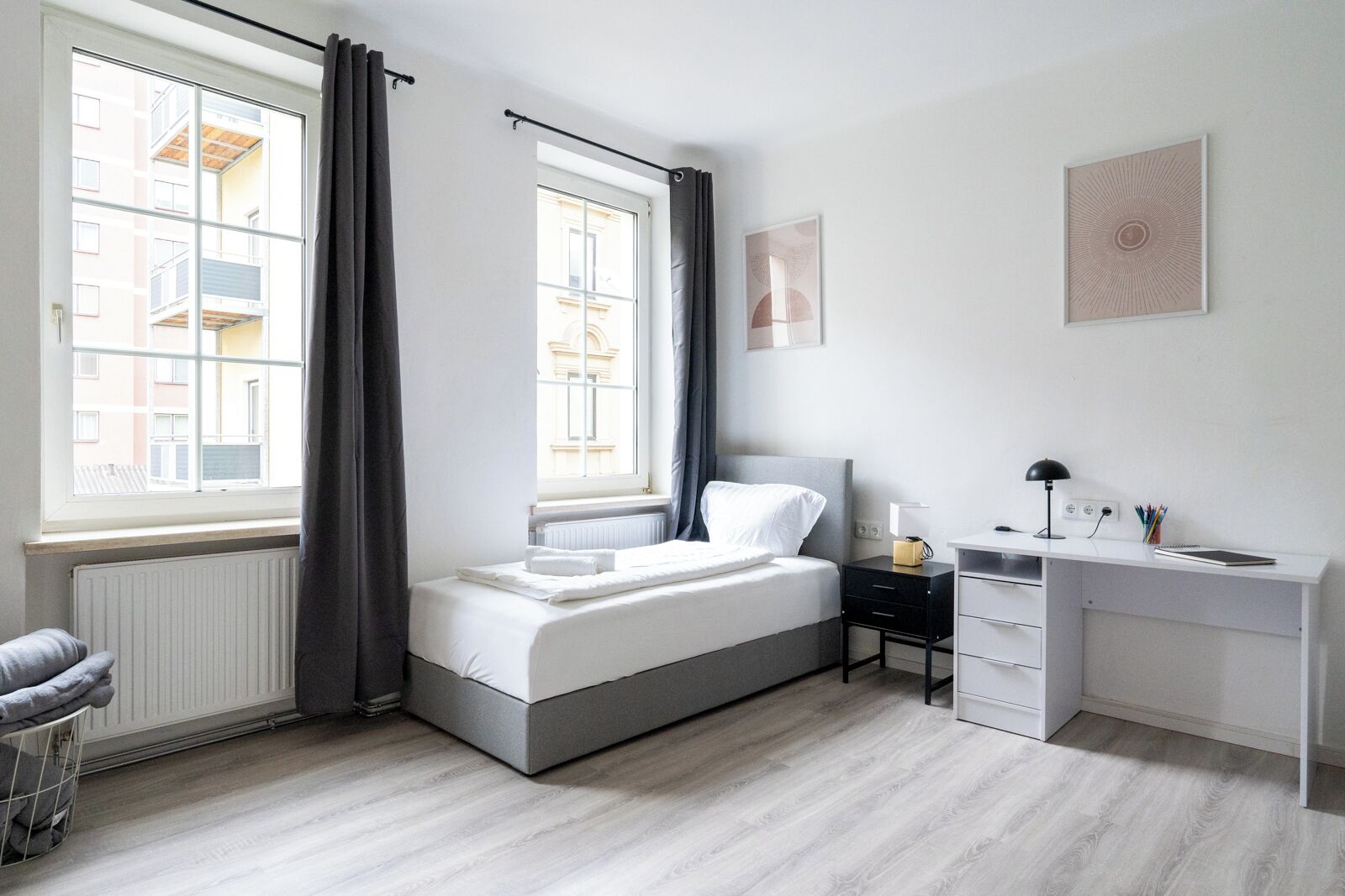 A bedroom with a bed, a desk, and a chair. There are two windows with curtains, a radiator, and a wall with a picture.