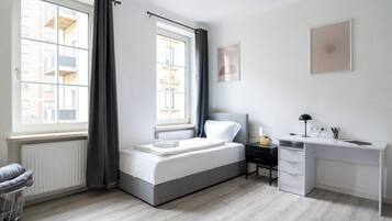 A bedroom with a bed, a desk, and a chair. There are two windows with curtains, a radiator, and a wall with a picture.