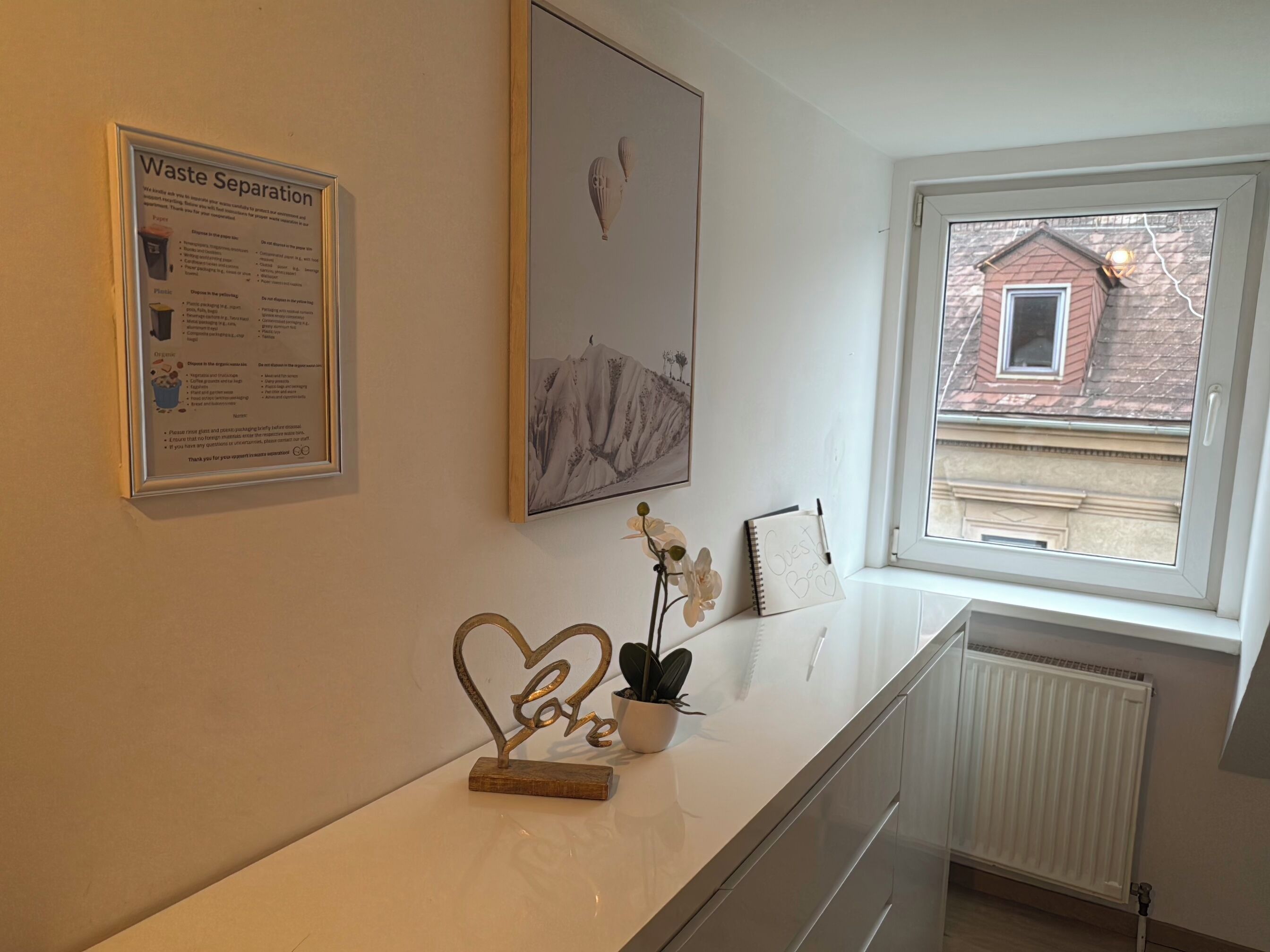 A room with a framed waste separation poster, a framed landscape painting, a window with a view of a building, a potted orchid, and a notebook on a counter.
