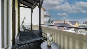 A balcony with a hanging chair, a small table with a vase, and a view of houses in the distance. - COY Apartments Leoben - Stay Comfortable & Work Smart (Leoben)