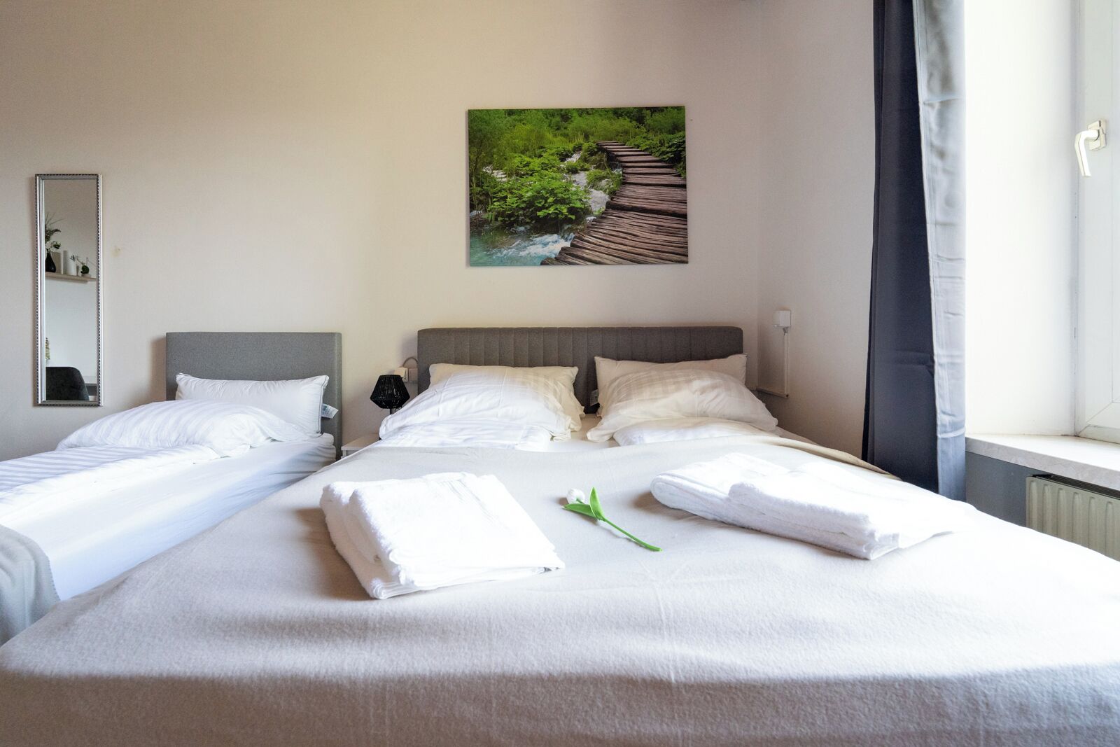Habitación de hotel con dos camas individuales, una cama grande con toallas, un cuadro de un sendero en el bosque, una ventana con cortinas y un radiador.
