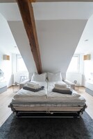 Ein Schlafzimmer im Loft-Stil mit hohen Decken, freiliegenden Holzbalken, einem großen Bett mit ordentlich zusammengefalteten Decken und Blick ins Freie durch ein Fenster.