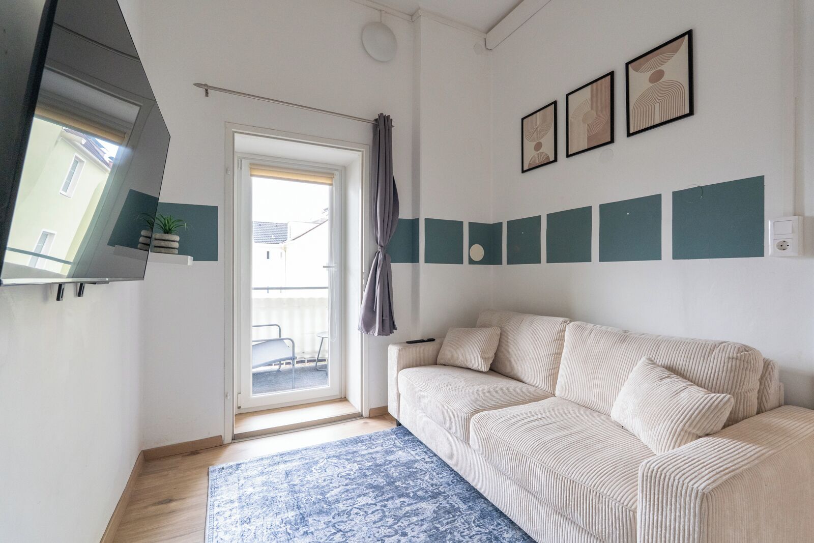 A modern living room with a sofa, a blue patterned rug, a sliding glass door, and a small balcony with a plant.