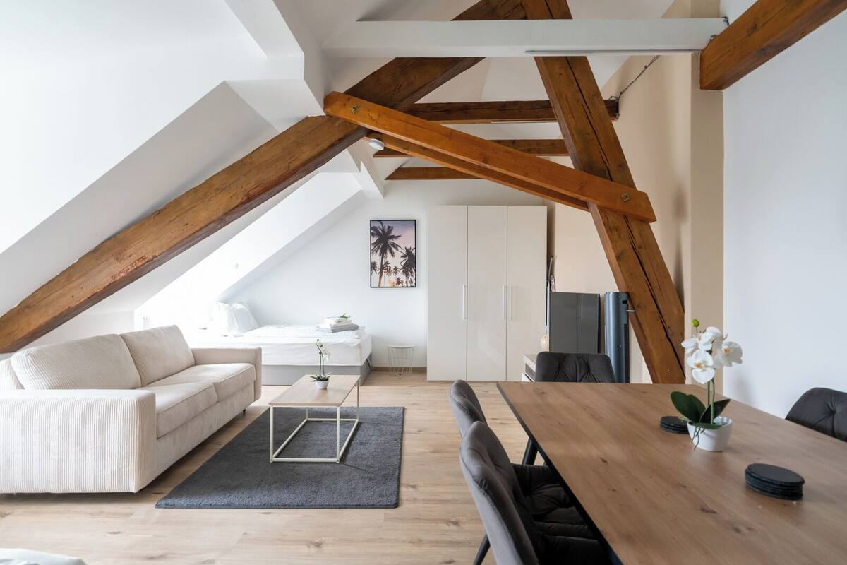 A modern loft bedroom with a sloped ceiling, a wooden dining table, a sofa, and a small coffee table.