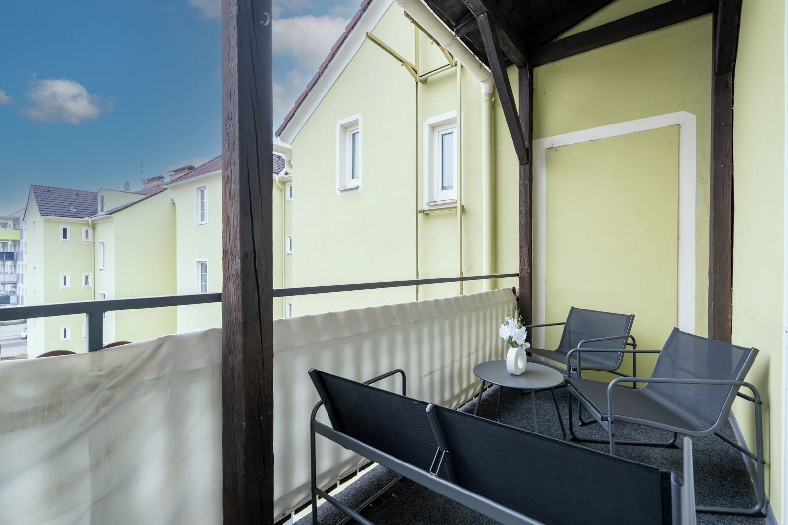 En balkon med to stole og et lille bord, med udsigt over en række gule bygninger.