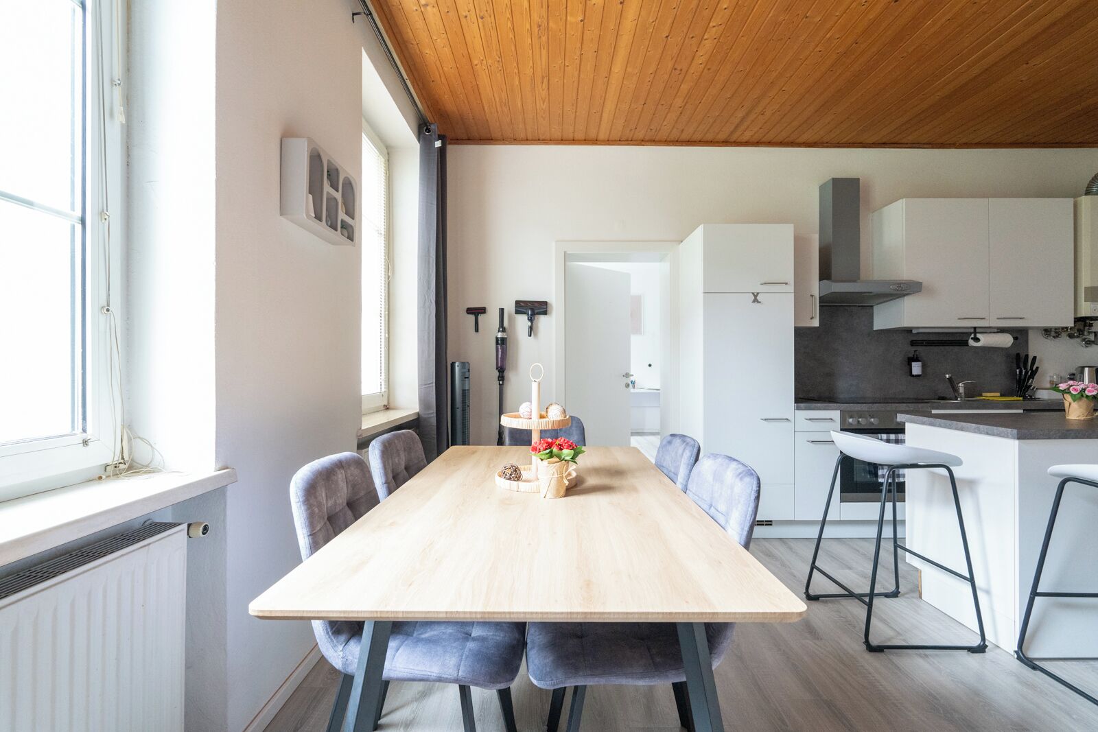 Eine moderne Küche mit weißen Schränken, einer Holzbalkendecke und einem Essbereich mit einem hellen Tisch und grauen Stühlen.