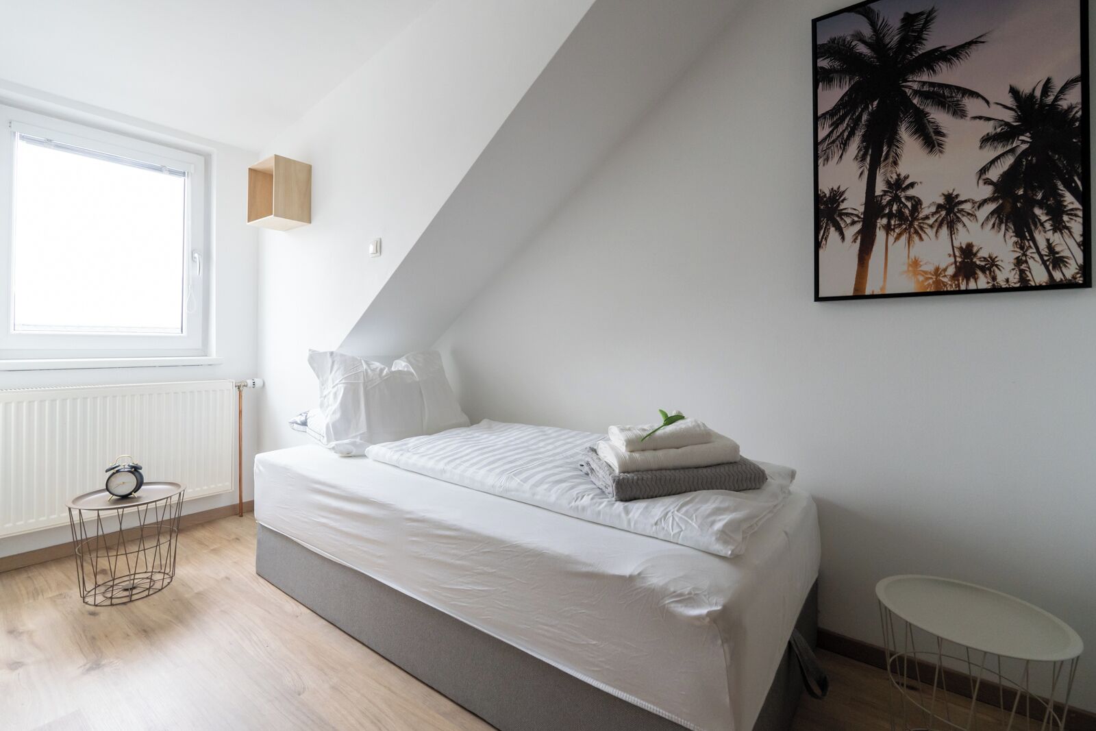 A bedroom with a bed, a window, a radiator, a small table, and a framed picture on the wall.
