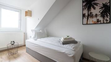 A bedroom with a bed, a window, a radiator, a small table, and a framed picture on the wall.