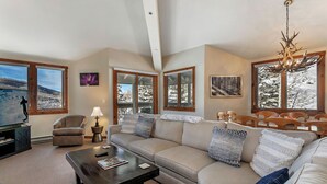 A spacious living room with a large sectional sofa, a coffee table, and a television. There are large windows offering views of a snowy landscape. - Beautiful Mountain Residence, Ski in/out, Year-Round Pool and Hot Tubs (Beaver Creek)