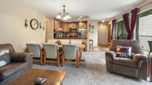 A living room with a brown leather sofa, a wooden dining table with four chairs, a coffee table, and a kitchen in the background.