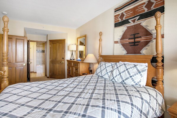 A bedroom with a wooden four-poster bed, a checkered bedspread, a mirror, and a wall with a geometric tapestry.