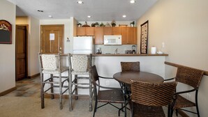 A compact kitchen with wooden cabinets, a microwave, and a refrigerator. There is a dining area with wicker chairs and a round table.