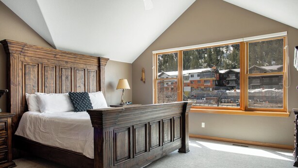 A bedroom with a large bed, a nightstand, a lamp, and a window with a view of buildings. - Chic Streamside Home, Hot Tub, Minutes from Resort (Avon)