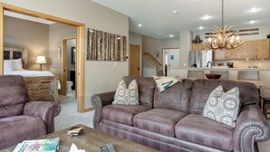A living room with a brown leather sofa, a wooden coffee table, and a dining area with a chandelier. - Chic Streamside Home, Hot Tub, Minutes from Resort (Avon)