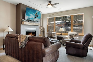 A living room with a fireplace, wall-mounted TV, and a large window. There are two brown sofas and two patterned armchairs arranged around a coffee table. A ceiling fan is on the white ceiling.