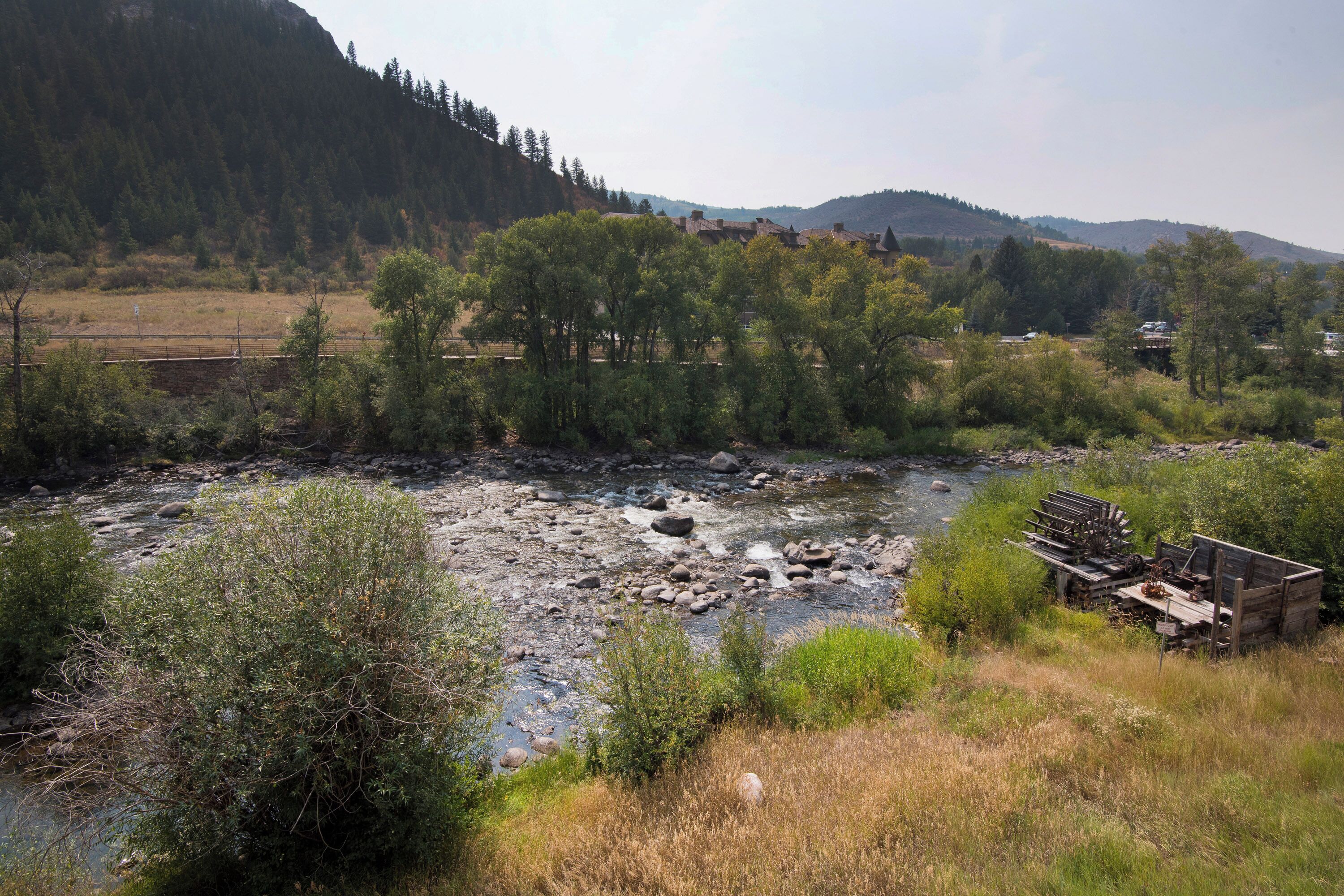 A river with a wooden structure in the foreground, surrounded by trees and hills in the background.