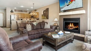 A living room with a fireplace, a television, and a kitchen in the background. - Chic Streamside Home, Hot Tub, Minutes from Resort (Avon)
