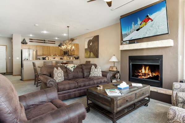 Una sala de estar con chimenea, televisor y cocina al fondo.