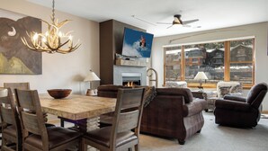 A cozy living room with a fireplace, a large window with a view, and a dining area with wooden chairs and a table. - Chic Streamside Home, Hot Tub, Minutes from Resort (Avon)