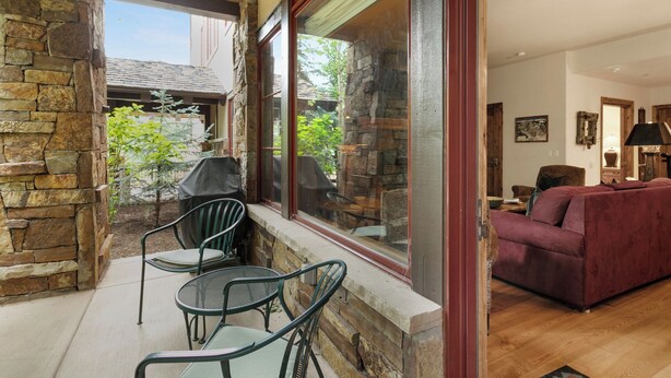 A stone exterior wall, a covered patio with chairs, a red sofa, and a view of a house through a large window.