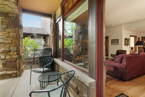 A stone exterior wall, a covered patio with chairs, a red sofa, and a view of a house through a large window. - Gorgeous Mountainside Condo, A+ Location, A/C, Ski in/Out Hot Tubs (Beaver Creek)